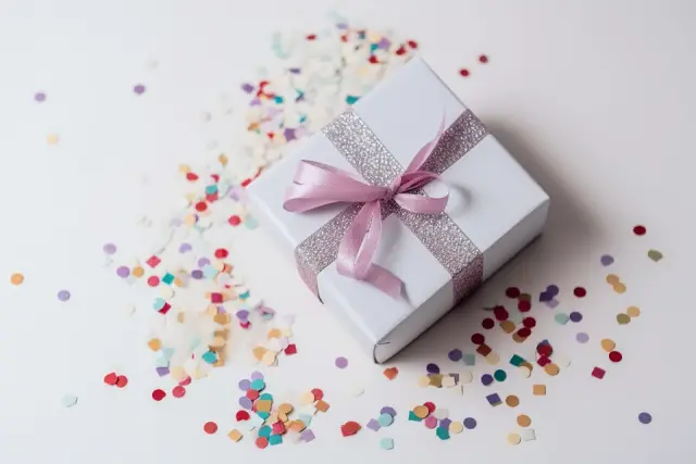 Un paquet cadeau blanc noué d'un ruban rose, entouré de confettis de couleur sur une table blanche