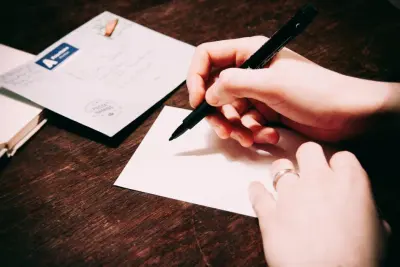 Deux mains écrivent au feutre noir sur une carte blanche posée sur une table en bois, à côté d'une enveloppe