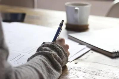 Une main en pull gris prend des notes au stylo sur une feuille, sur une table en bois près d'une tasse