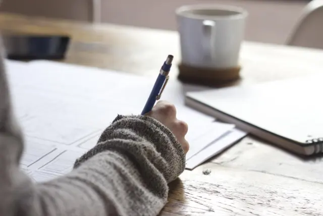 Une main en pull gris prend des notes au stylo sur une feuille, sur une table en bois près d'une tasse