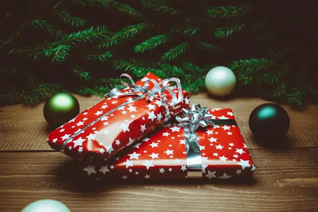 Deux paquets cadeaux en papier rouge à étoiles blanches posés sur un parquet, sous des branches de sapin et des boules vertes